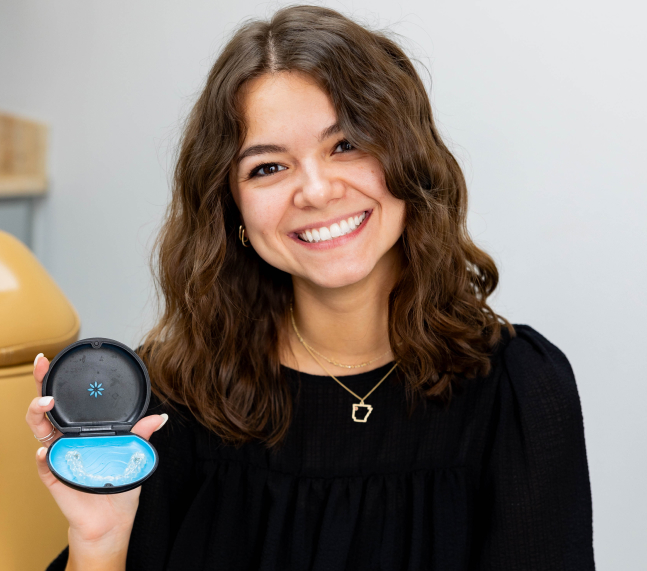 Smiling woman holding blue dental aligner case in orthodontic office setting.